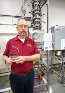 A man in a red shirt holding a glass award stands in a lab