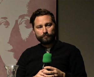 A bearded man talks into a microphone with a green foam head