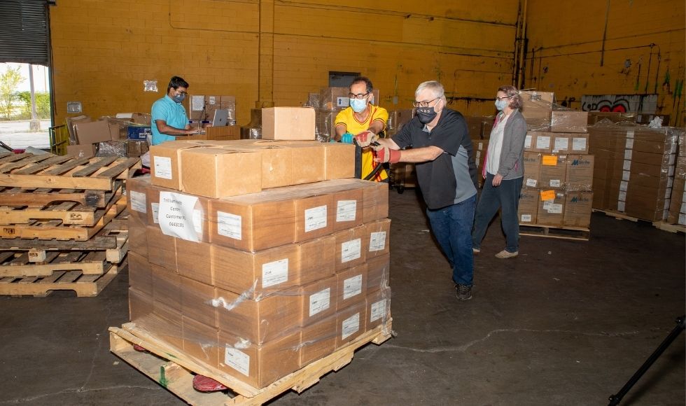 4 people, two of whom are pushing a pallet loaded with cardboard boxes.