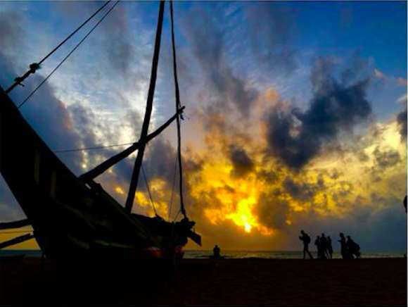 An image of a setting sun slipping behind the clouds. The photo is taken from a beach and a silhouette of a boat and a family are in the foreground. 
