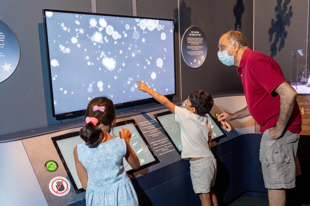 A photo of a man and two children engaging with a screen and infographics. All three people are masked. 