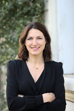 A photo of Tracey Dobbin pictured from the waist up with her arms crossed in front of her and smiling at the camera. She is wearing a black shirt and there is some greenery out of focus behind her. 