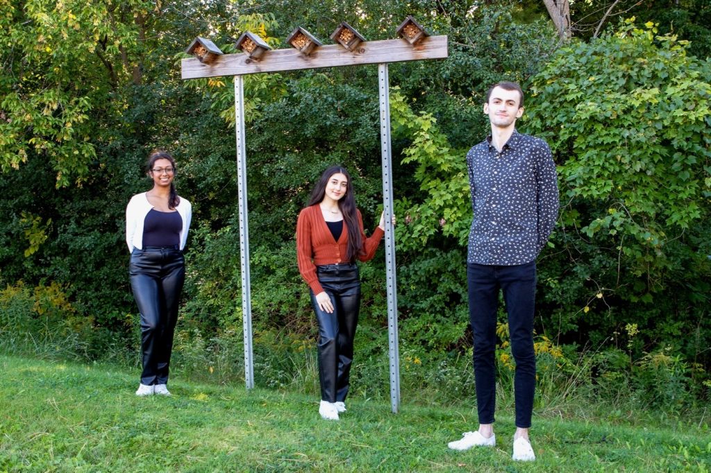 A photo of three McMaster students standing outdoors and smiling at the cmaera. They are standing in front of a pair of posts sticking out of the ground that are holding up a beam of wood. Five bee houses are sitting on top of the beam of wood. 