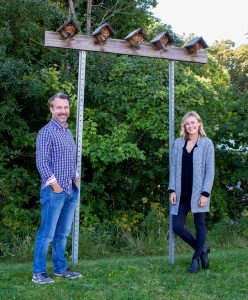 A photo of a man and woman standing outdoors smiling at the camera. They are standing in front of a pair of posts sticking out of the ground that are holding up a beam of wood. Five bee houses sit on top of the beam of wood. 