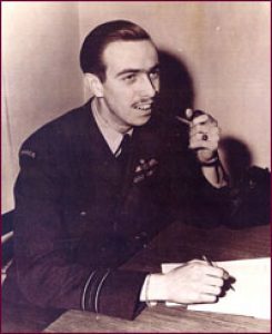 A black and white photo of a man in a WWII pilot's uniform. The man has a mustache and a pipe, and is writing on a piece of paper on the table in front of him 