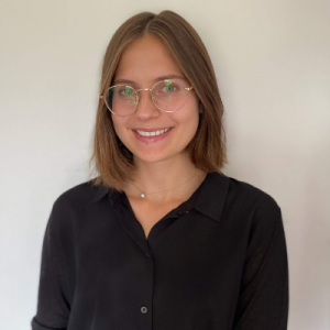 A photo of Zuzanna smiling at the camera. She is wearing a black button-up shirt and round, gold-rimmed glasses. 