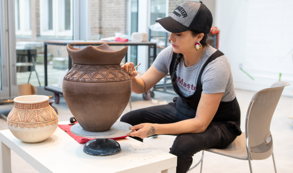 Santee Smith carving a brown clay pot. She is wearing large earrings, a grey t-shirt, black overalls and a black and grey baseball cap. She is seated on the edge of a chair, working at a table.
