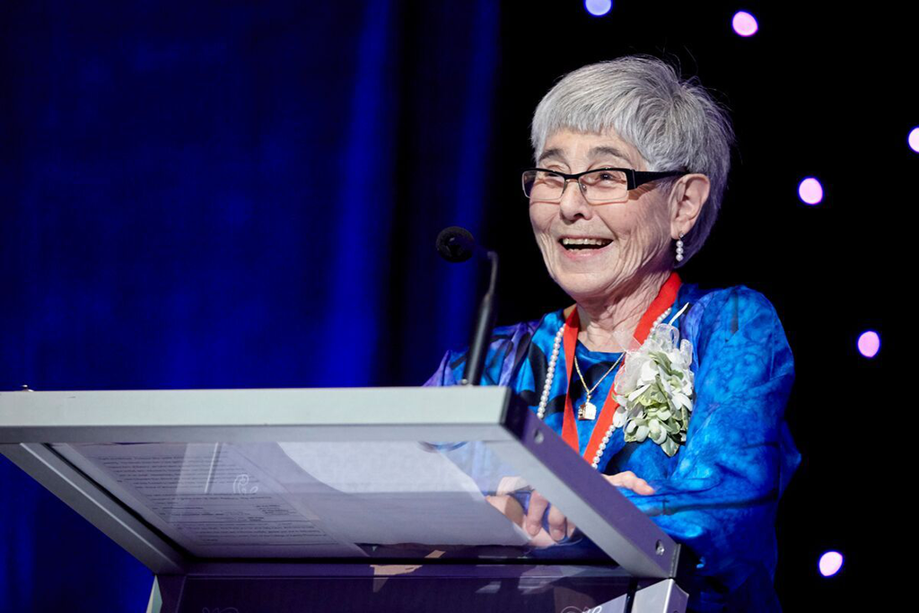 A photo of May Cohen standing at a podium. She is smiling and wearing a blue dress and a corsage on her lapel. 