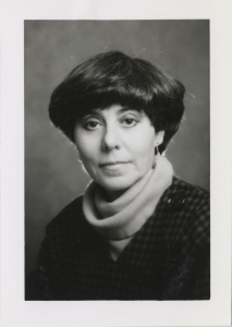 A black-and-white photo of May Cohen. She is wearing a turtleneck and sweater and is looking at the camera without smiling. 