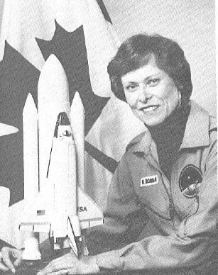 A black-and-white photo of Robert Bondar wearing a space jumpsuit. She is sitting in front of the Canadian flag and has her arms around a model of the space shuttle Discovery.