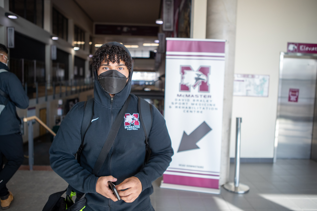 A masked McMaster student stares straight at the camera. A banner advertising the David Braley Athletics Centre (DBAC) is behind him over his shoulder. 