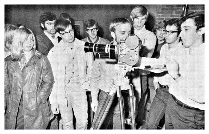 A group of students at McMaster surround a camera as one student on the far right directs the scene. Black and white image.