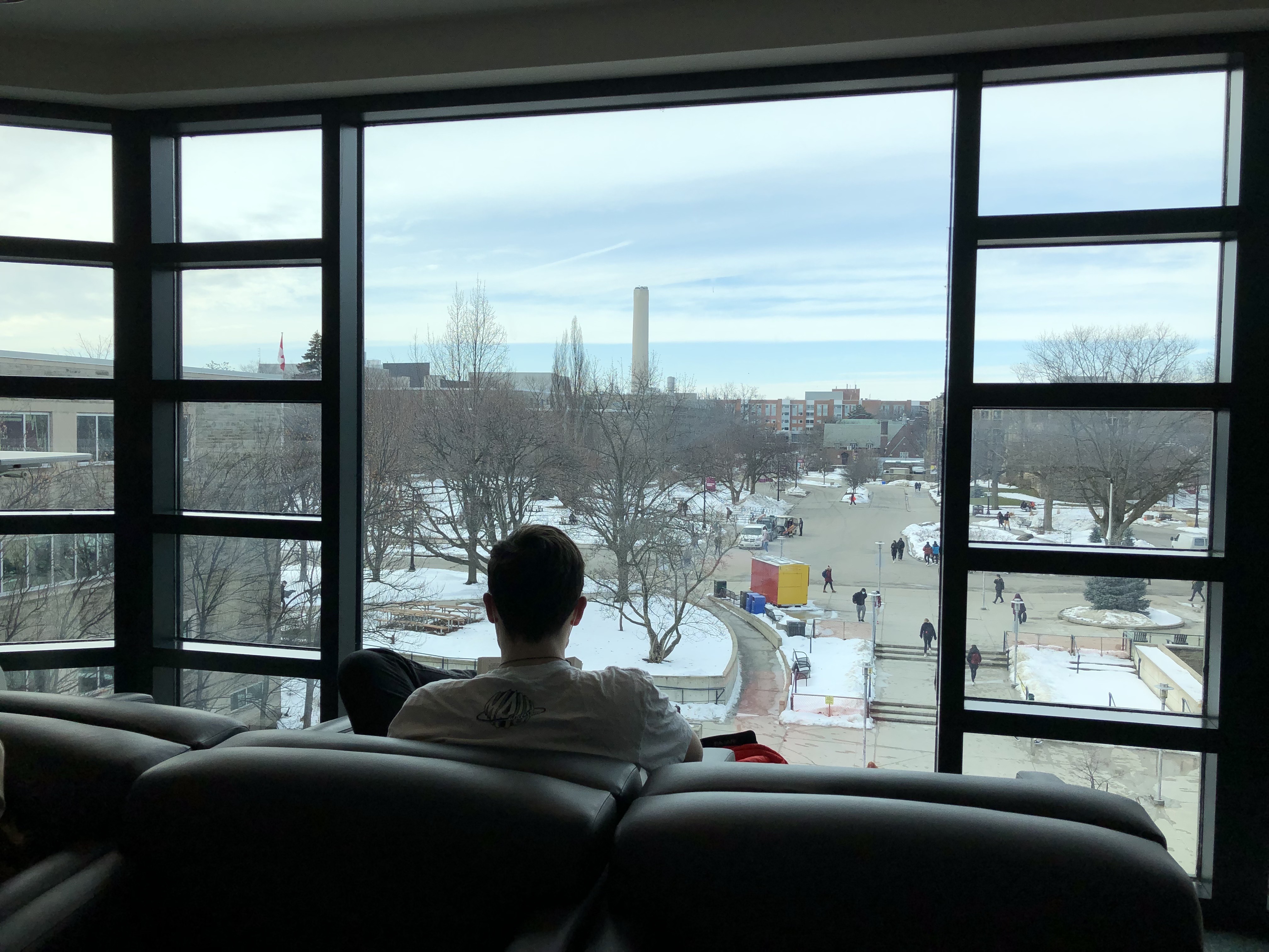 A photo of a student sitting on a couch reading in front of a big window that provides a view of McMaster's campus 