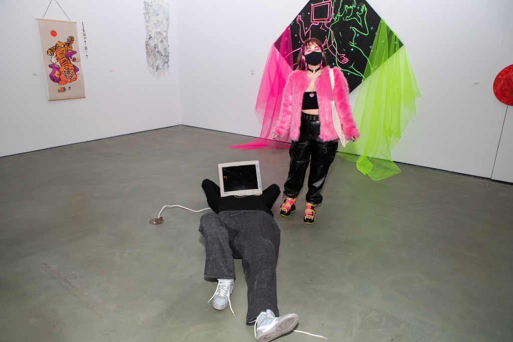A person with a mock computer screen over their head lies on the floor. PauTheRebel stands behind them. Behind PauTheRebel is a black and neon pink and neon green painting. 