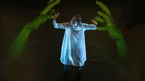 A person with their arms stretched over their head. Behind them is a screen with two green hands. 