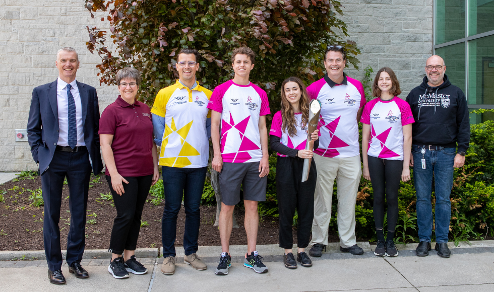 A group of researchers from McMaster and University of Birmingham gather outside of a McMaster building on a sunny day. Four are wearing relay shirts and the rest are wearing business attire and McMaster gear