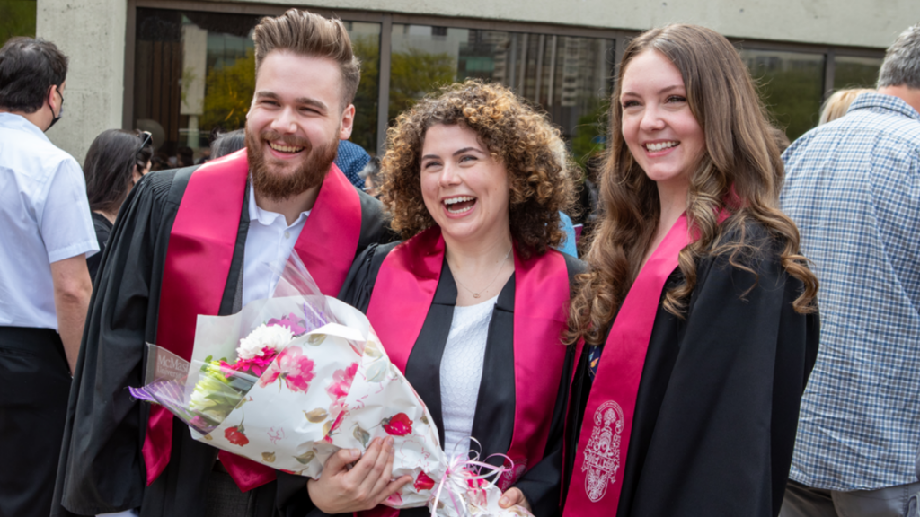 Three smiling McMaster graduates pose for a photo. One is holding a bouquet of flowers