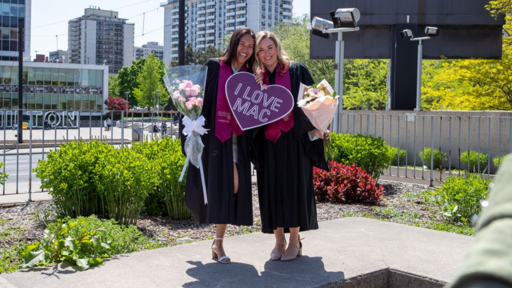Two graduates pose for a photo while holding a sign that says 'I love Mac'