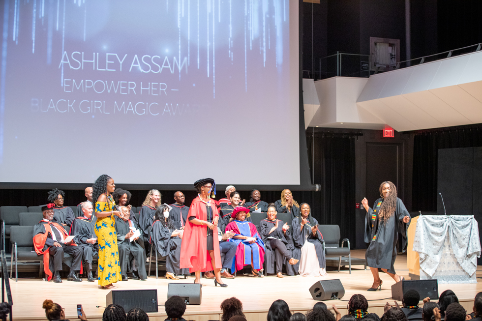 Ashley in her grad gown dances on stage, watched by the platform party as a screen behind them reads: Ashley Assam: Empower Her Black Girl Magic Award.