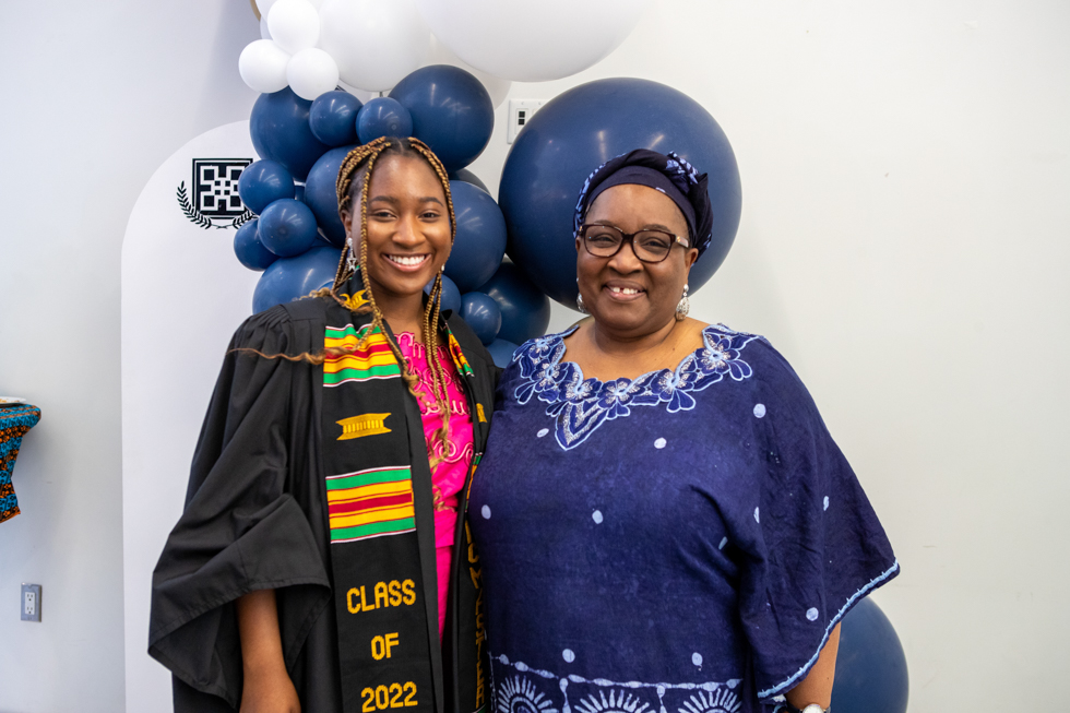 Kemi in her graduation gown and Kente stole, alongside her smiling mother, against a backdrop of balloons.