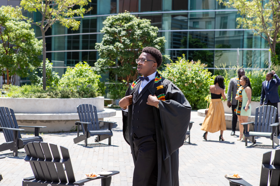 Cheikh in his bowtie, suit, grad gown and Kente Stole, walks through the LR Wilson courtyard in the sunshine