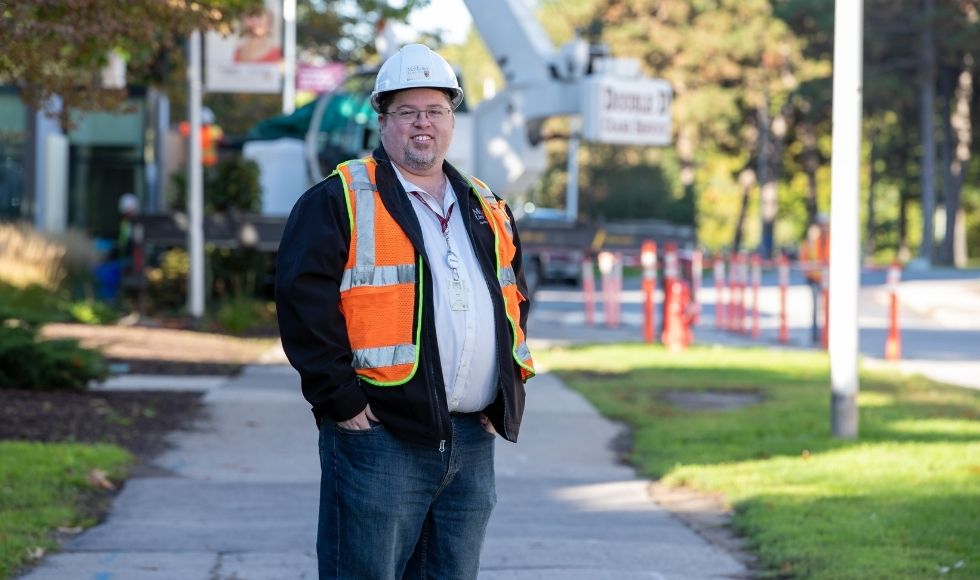 Wade, in a safety vest and hardhat, standing on a sidewalk with a bucket truck in the background.