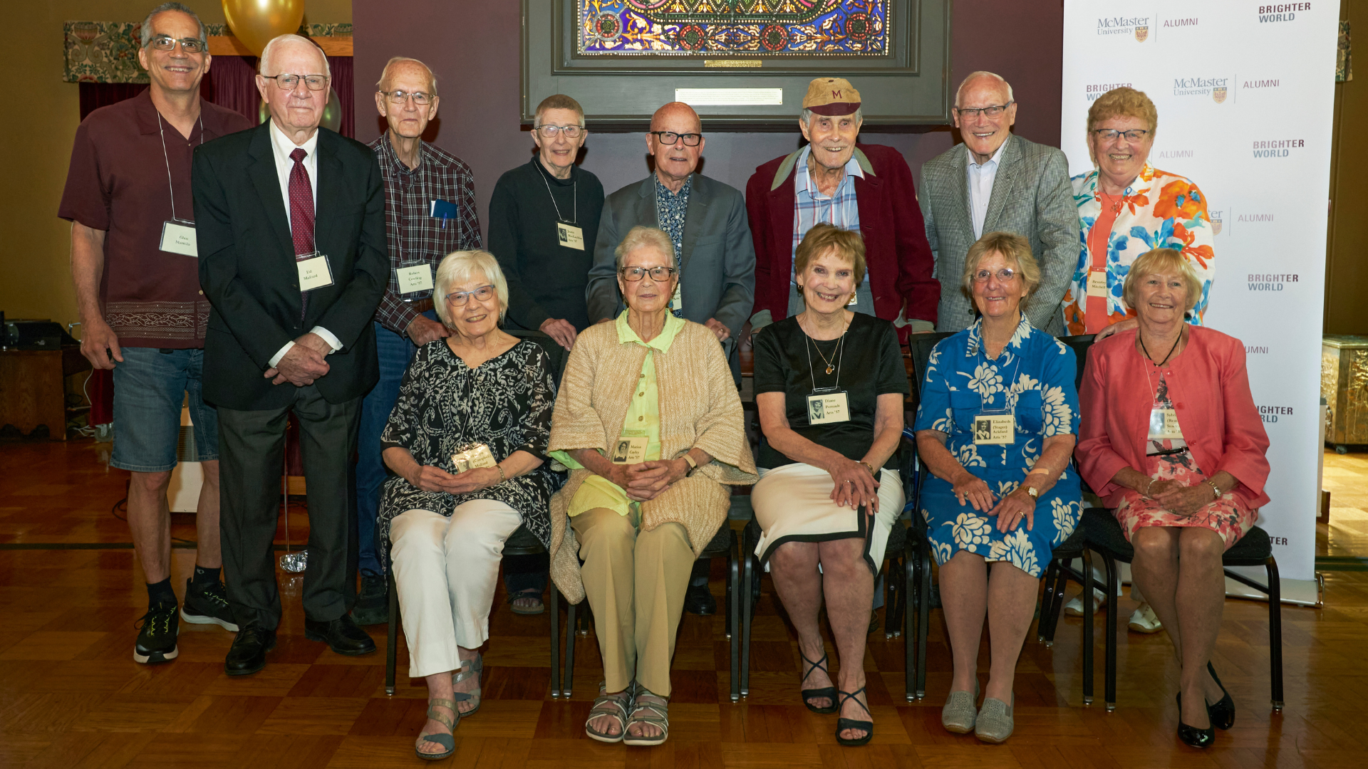 Thirteen McMaster alumni posing for a photo inside McMaster Alumni Hall. Some people are standing and some are seated. 