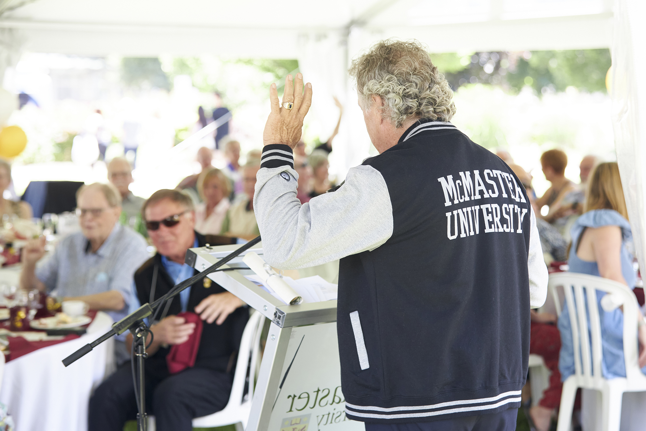 A man with his back turned to the camera delivers a speech to a crowd seated underneath a tent. The back of his jacket reads 'McMaster University.'