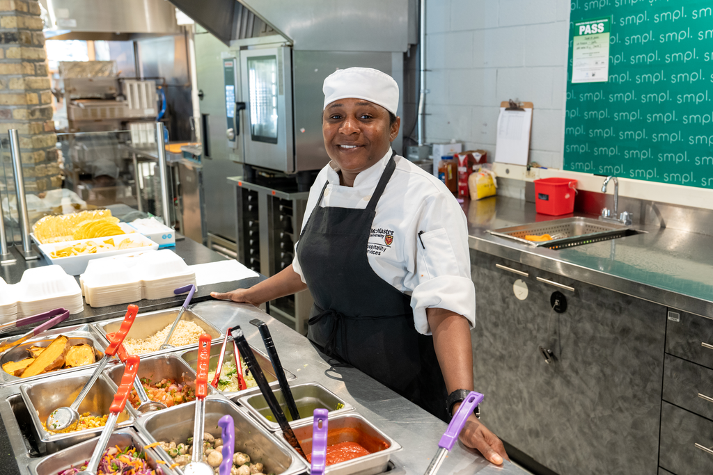 Marcia Traynor wearing chef's whites and standing behind a food serving station in McMaster's La Piazza. 