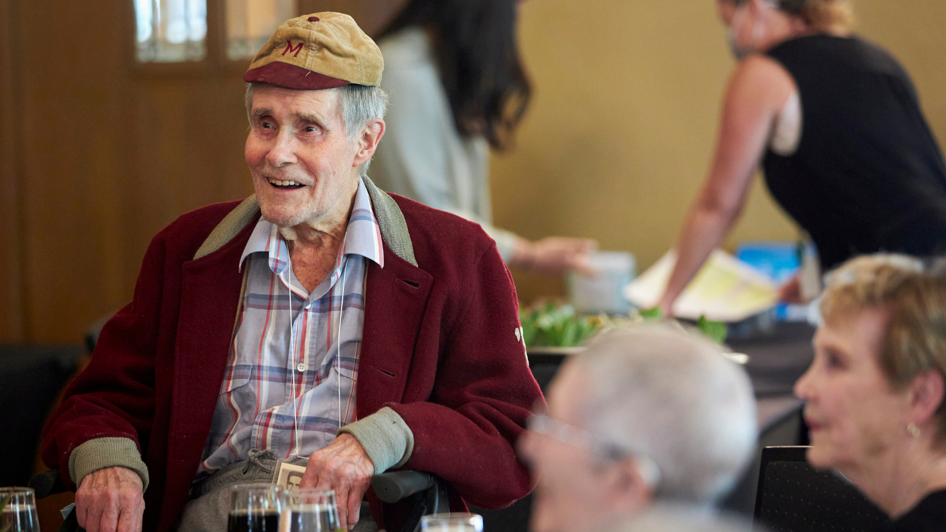 A McMaster graduate wearing a maroon McMaster jacket and maroon and beige hat smiles as he sits listening to speeches during a McMaster alumni luncheon
