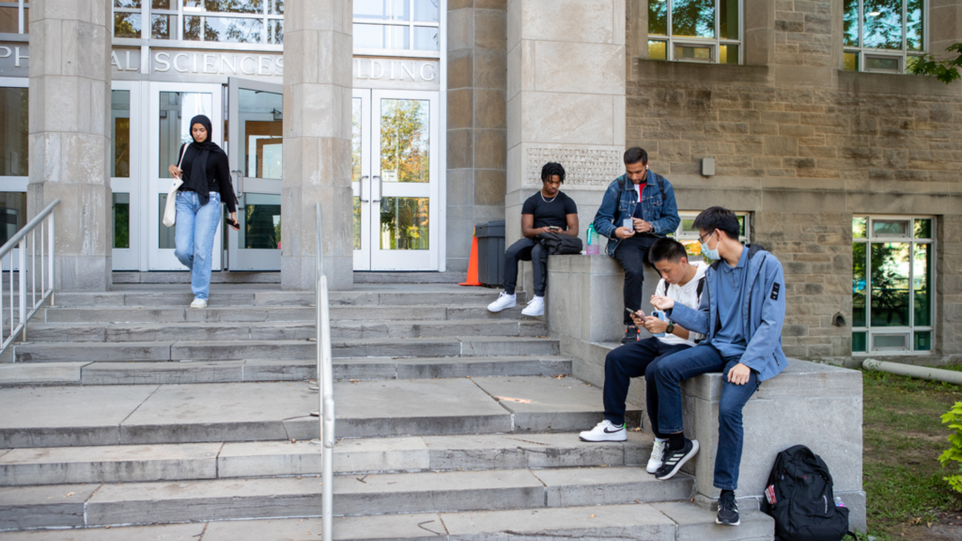 Four students seated looking at their phones as they sit outside the Burke Science Building. Another student is walking down the stairs to the building's entrance