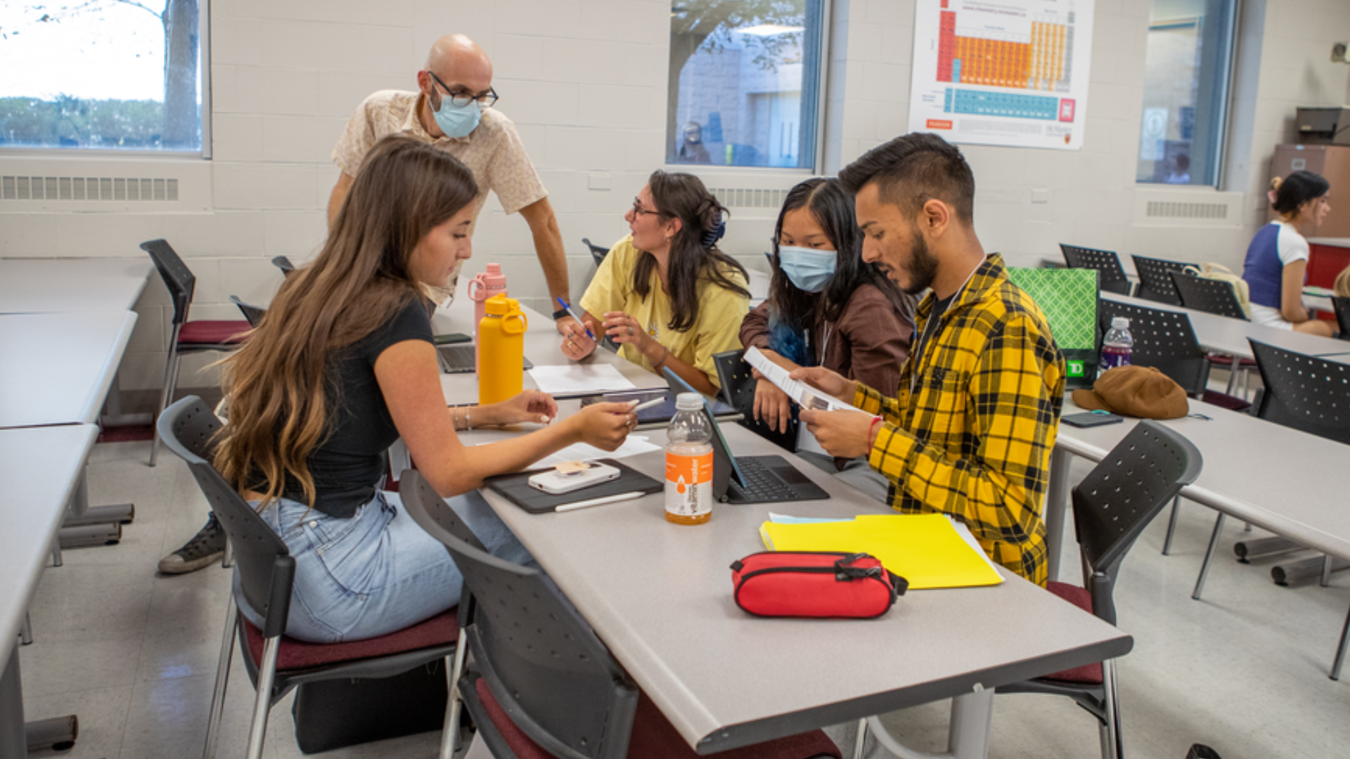 Four students seated at a table working together while their professor looks on