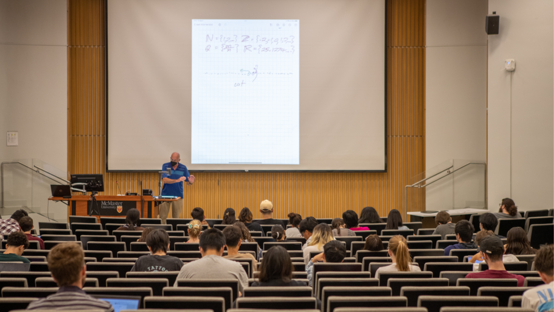 Students seated in a lecture hall while an instructor teaches. A document is projected up onto a screen at the front of the room. 