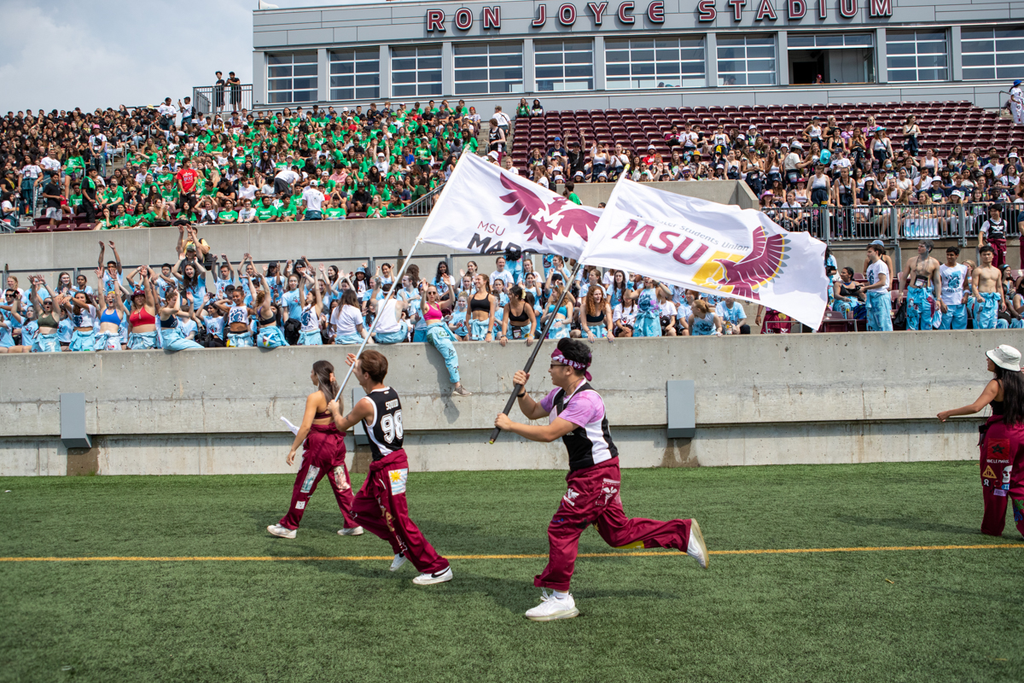Students running with large flags in front of a crowd at McMaster's Ron Joyce Stadium 