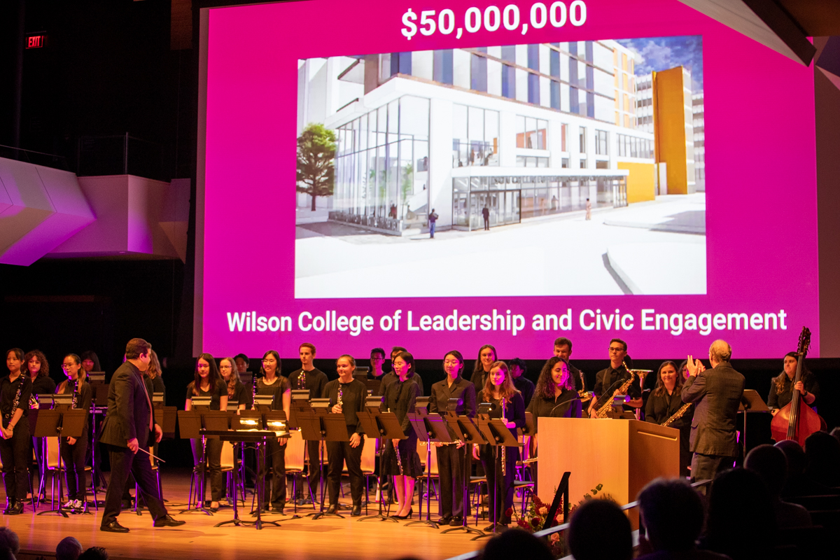 The McMaster Concert Band performs on stage. Behind them is a screen with a rendering of a building on it. 