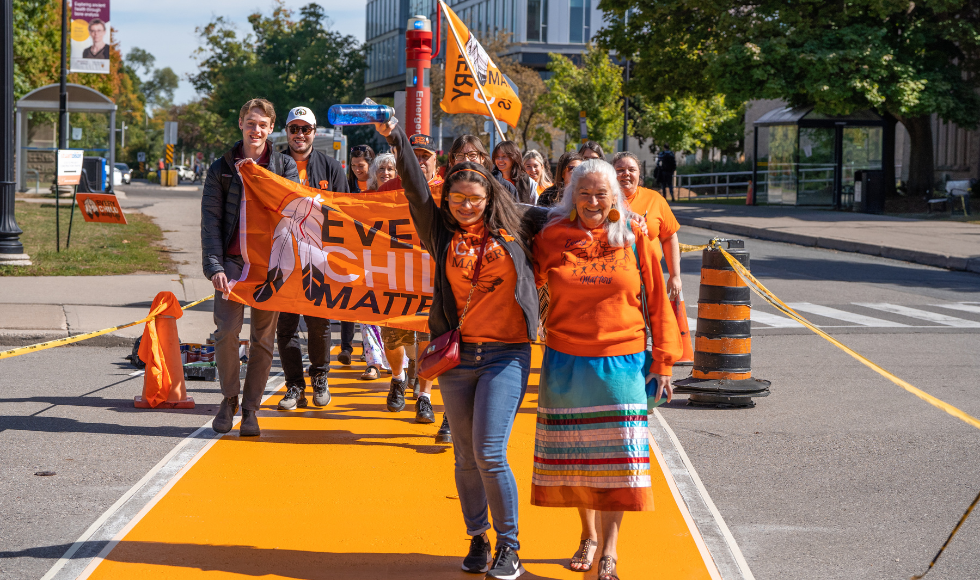 Led by a student and an elder, a group of people in orange shirts with an Every Child Matters flag cross a newly painted orange crosswalk.