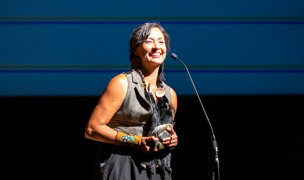A woman in a grey outfit stands on stage, smiling, addressing a crowd on a podium mic