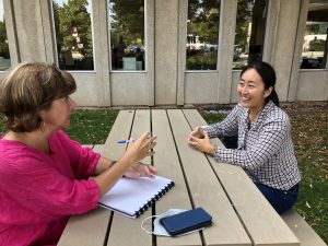 Avis Favaro and Katrina Choe engaged in conversation as they sit on opposite sides of a picnic bench outdoors. 