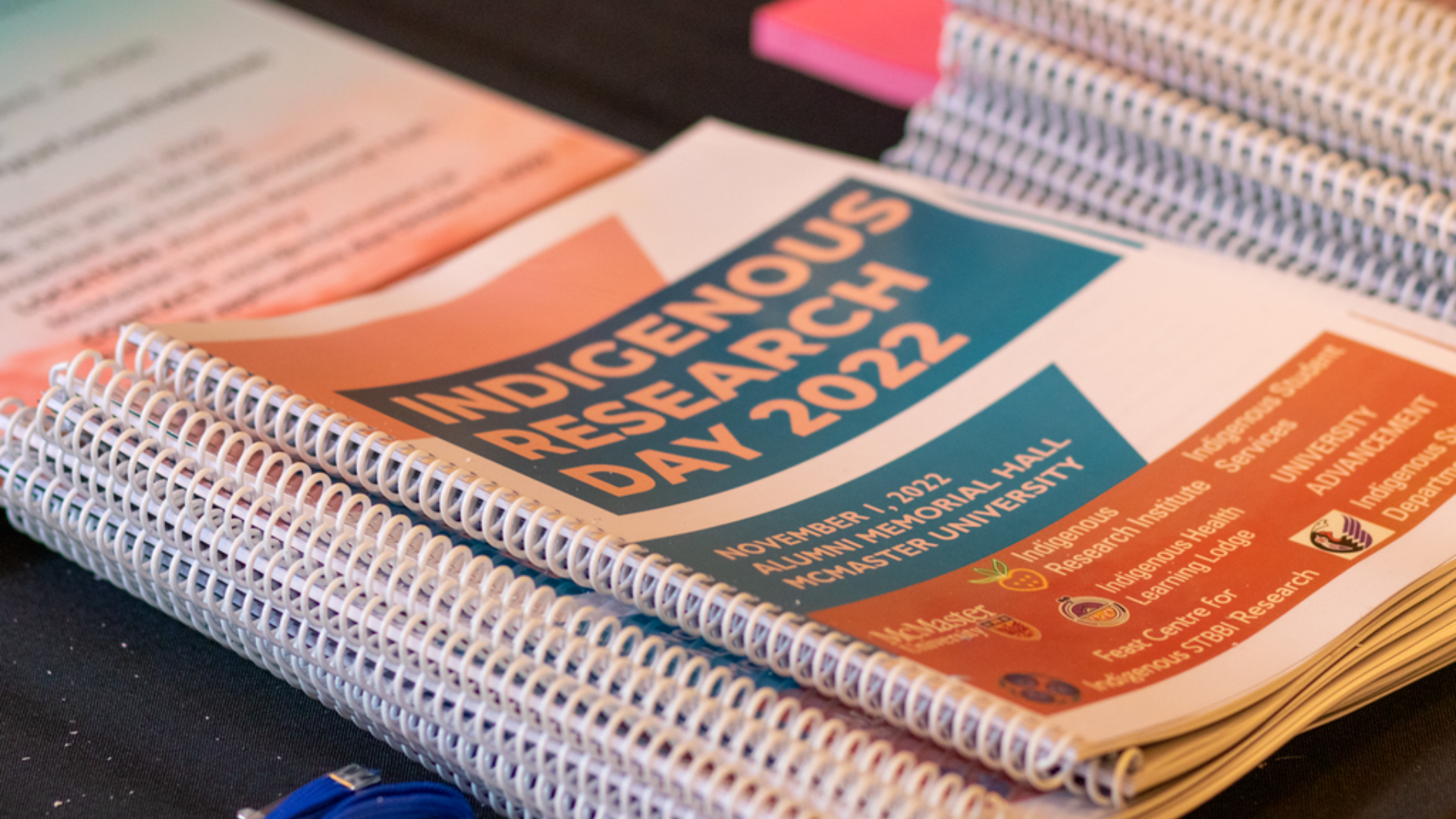 A pile of coil-bound notebooks sitting on a table. The cover of the notebook reads 'Indigenous Research Day 2022'