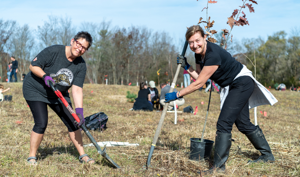 Two people with shovels look up and smile while digging into the ground outdoors.