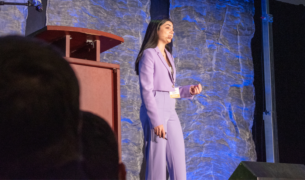 A person in a lilac suit with a microphone talking on stage.