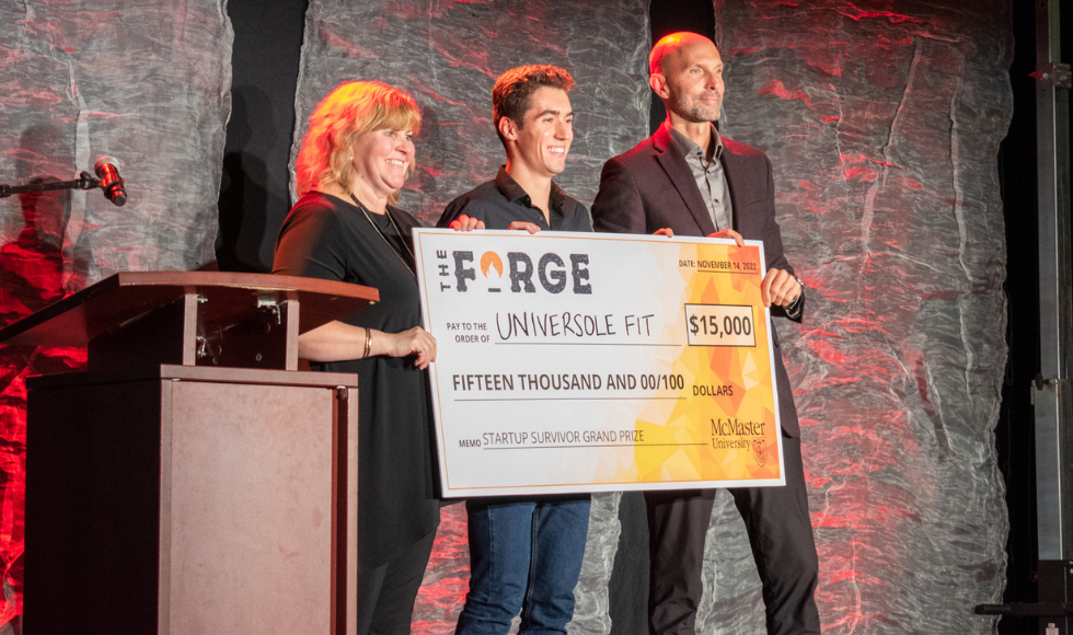 Provost Susan Tighe and Dean of Students Sean Van Koughnett stand on either side of a student holding an oversized novelty cheque for $15,000 on a stage.