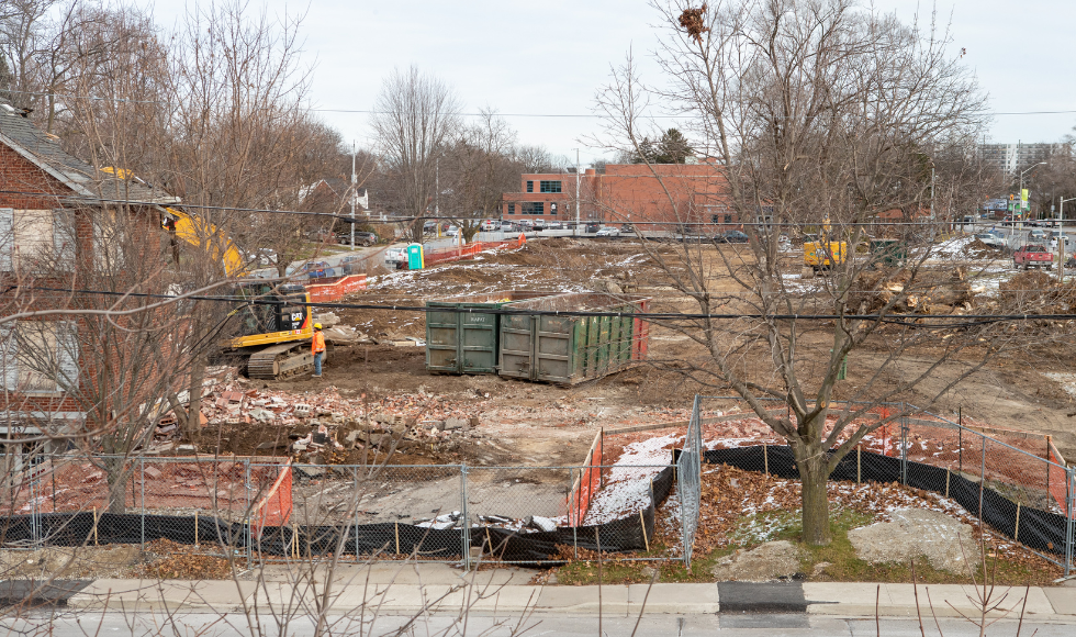 A construction zone with large metal disposal bins, heavy machinery and a house with boarded up windows.