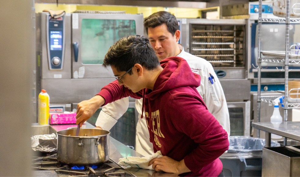 Two people in a kitchen. One of them is wearing a hoodie and is bent over a pot on the stove. The other is wearing a chef's jacket and standing back watching.