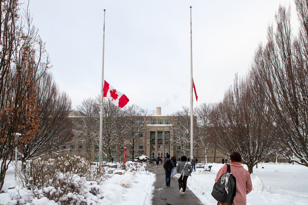 Two flags flying at half mast in front of a large building. There are students walking in the foreground of the photo. 