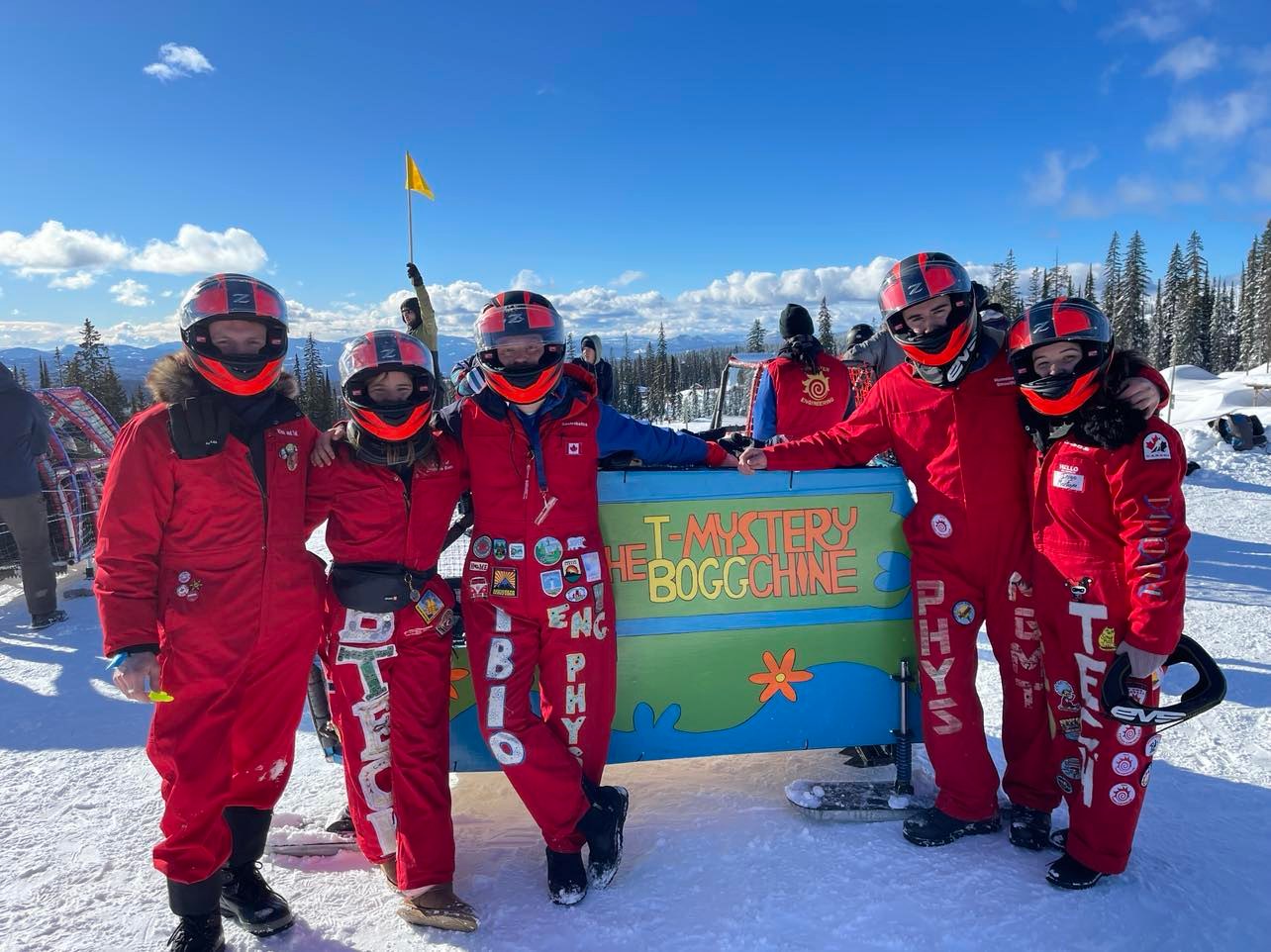 Five people in red snowsuits posing for a photo around a blue and green five-seater toboggan
