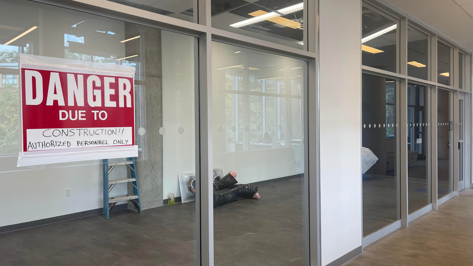 A row of floor-to-ceiling windows into a room that's mostly empty. On one of the windows there is a red and white sign reading, 'Danger due to construction!! Authorized personnel only.'