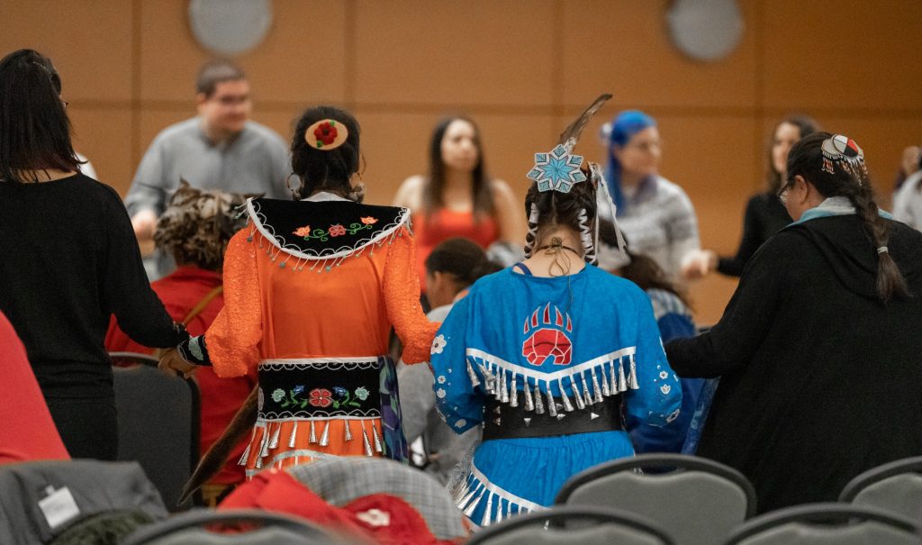 Dancers seen from behind holding hands during a powwow in CIBC hall.