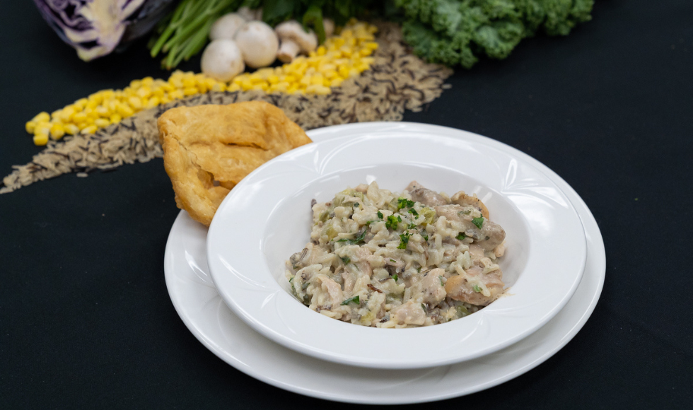 A white bowl of wild rice and chicken casserole on a white plate beside a piece of bannock, on a black tablecloth