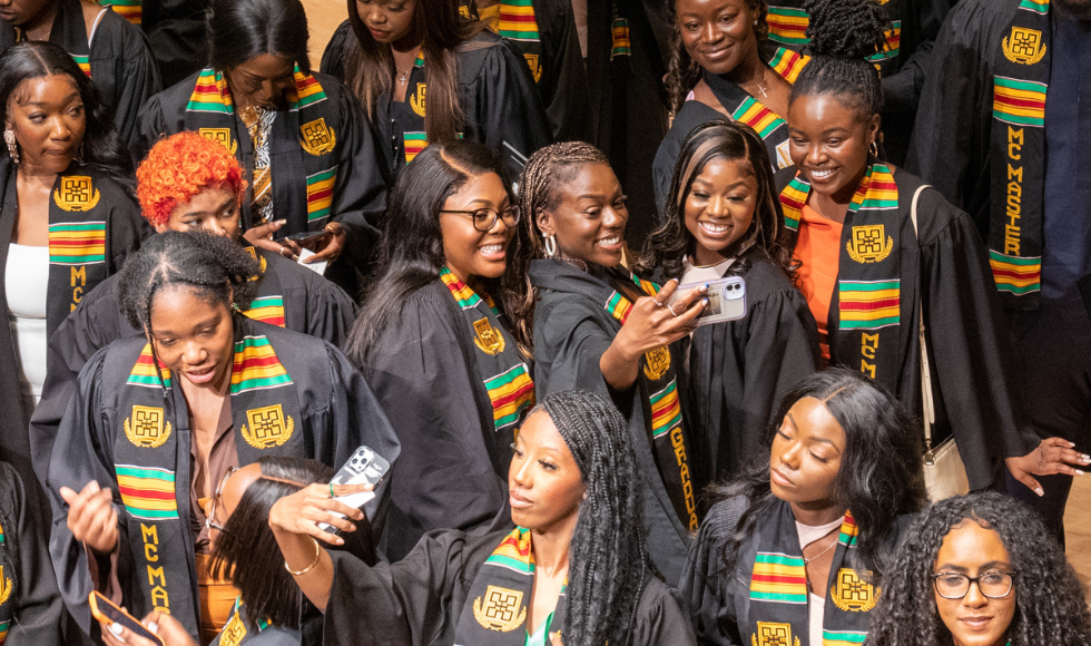A big group of students in their gowns and stoles, taking selfies and smiling, at the Black Excellence Graduation celebration.
